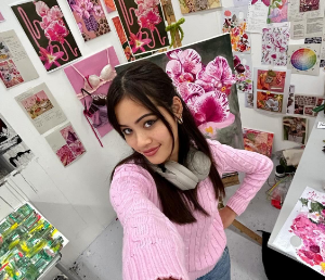 Nisha stands in her workspace surrounded by pink colourful paintings